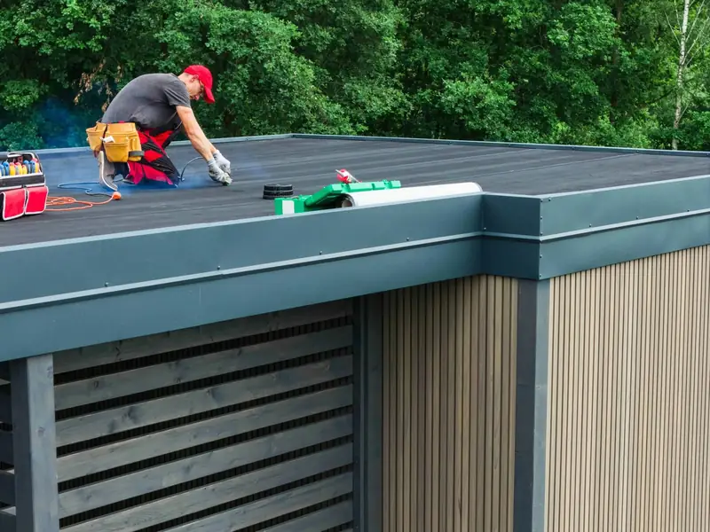 Roofer laying a membrane on the flat roof of a timber-clad garden building